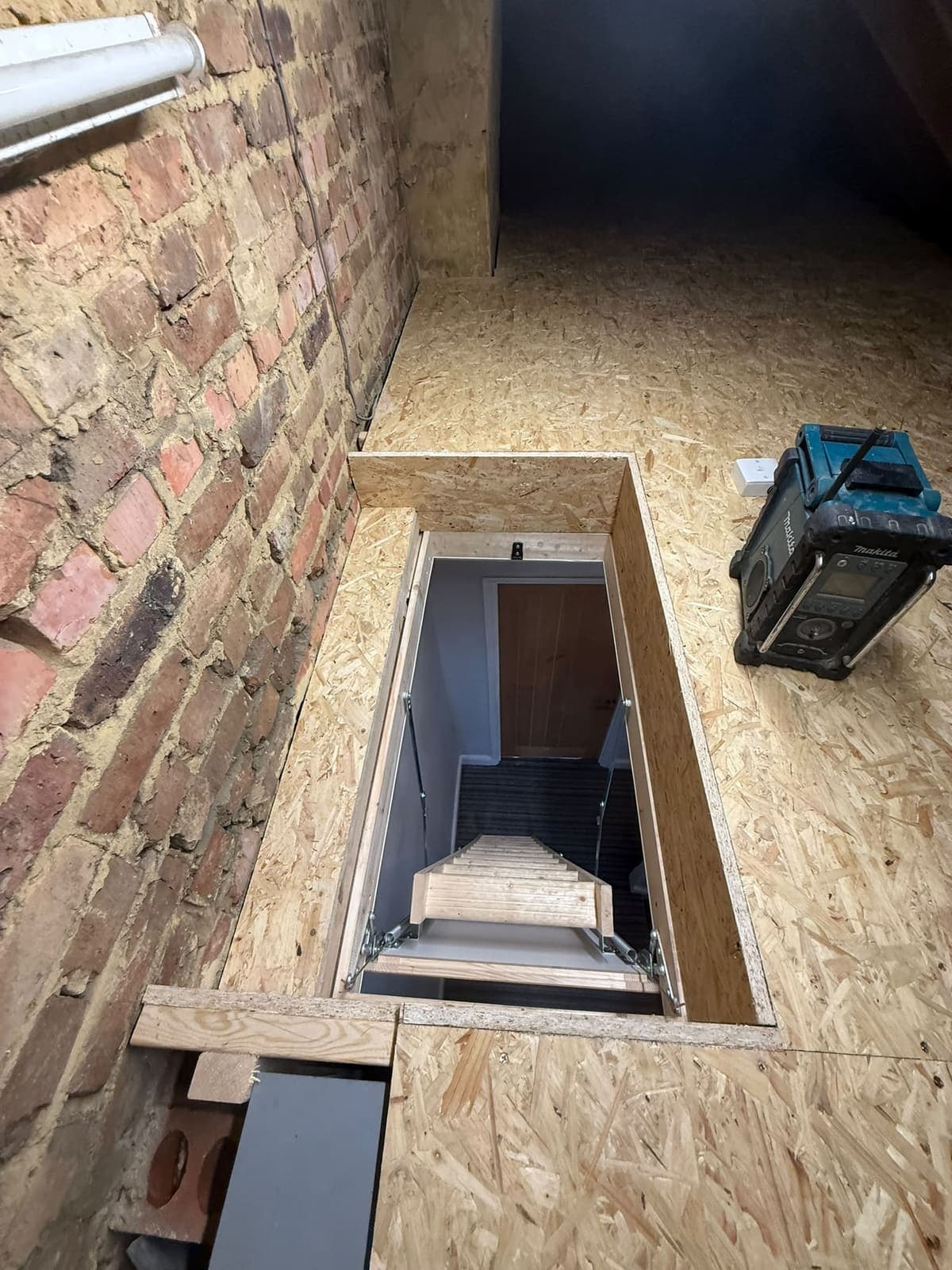 Lined loft hatch and ladder seen from above, beside the brick party wall